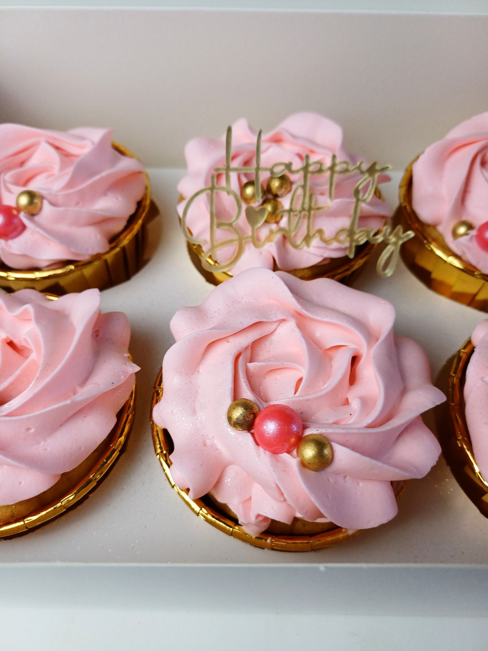 A box of six pink-frosted cupcakes decorated with gold and pink edible pearls, one featuring a gold 