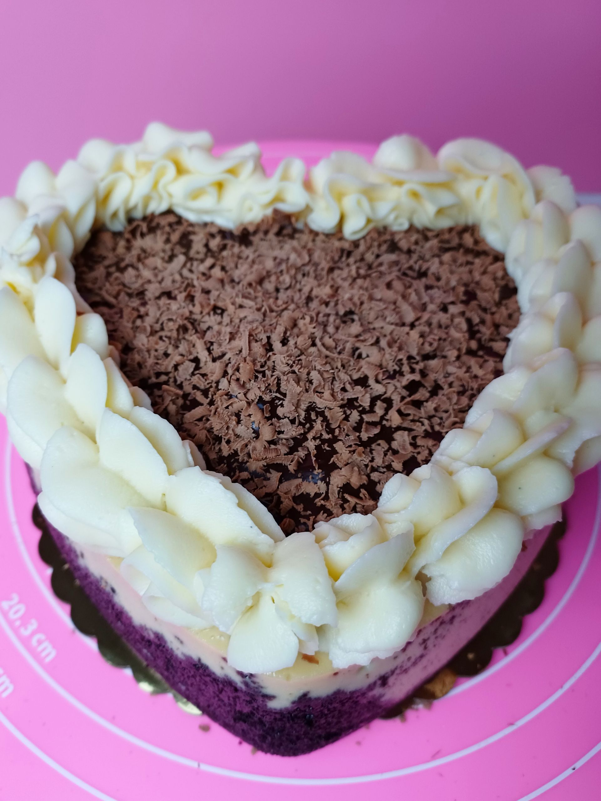 A heart-shaped cake with a dark purple base, a cream layer, chocolate shavings on top, and a white piped frosting border.