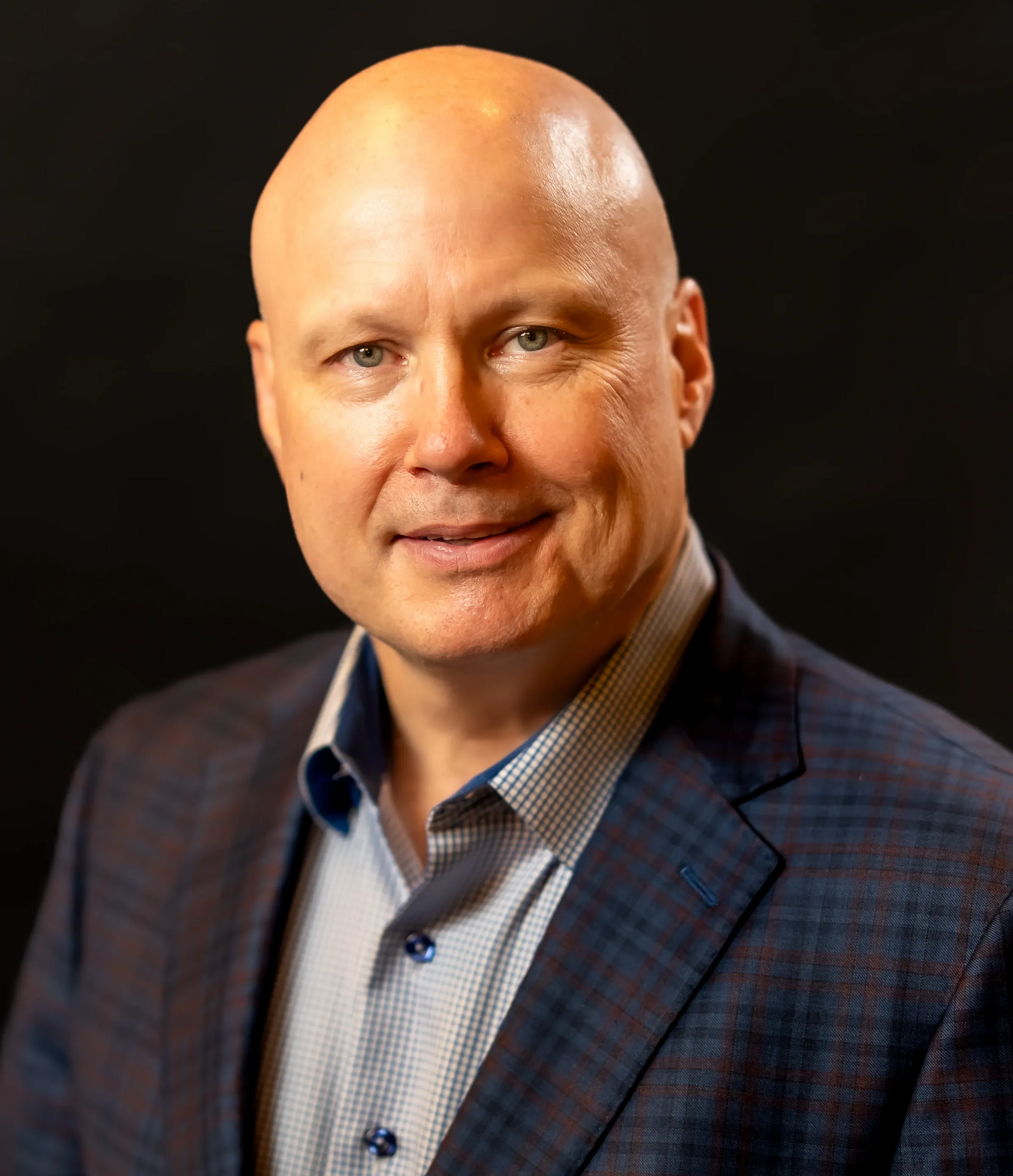 Bald man in a black suit with a gold tie, smiling, against a light gray wall.