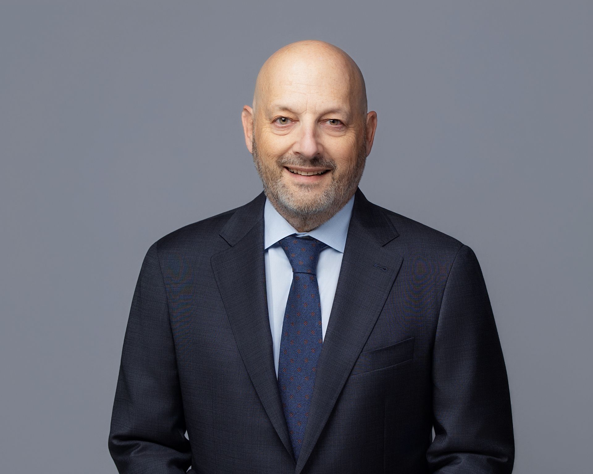 Man in suit smiling, with dark hair, on a gray backdrop.