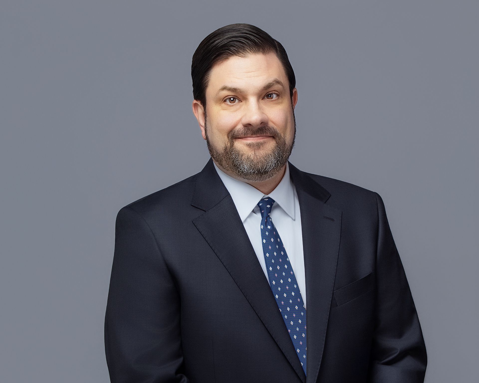 Man in navy suit and tie, smiling, posed against a gray backdrop.