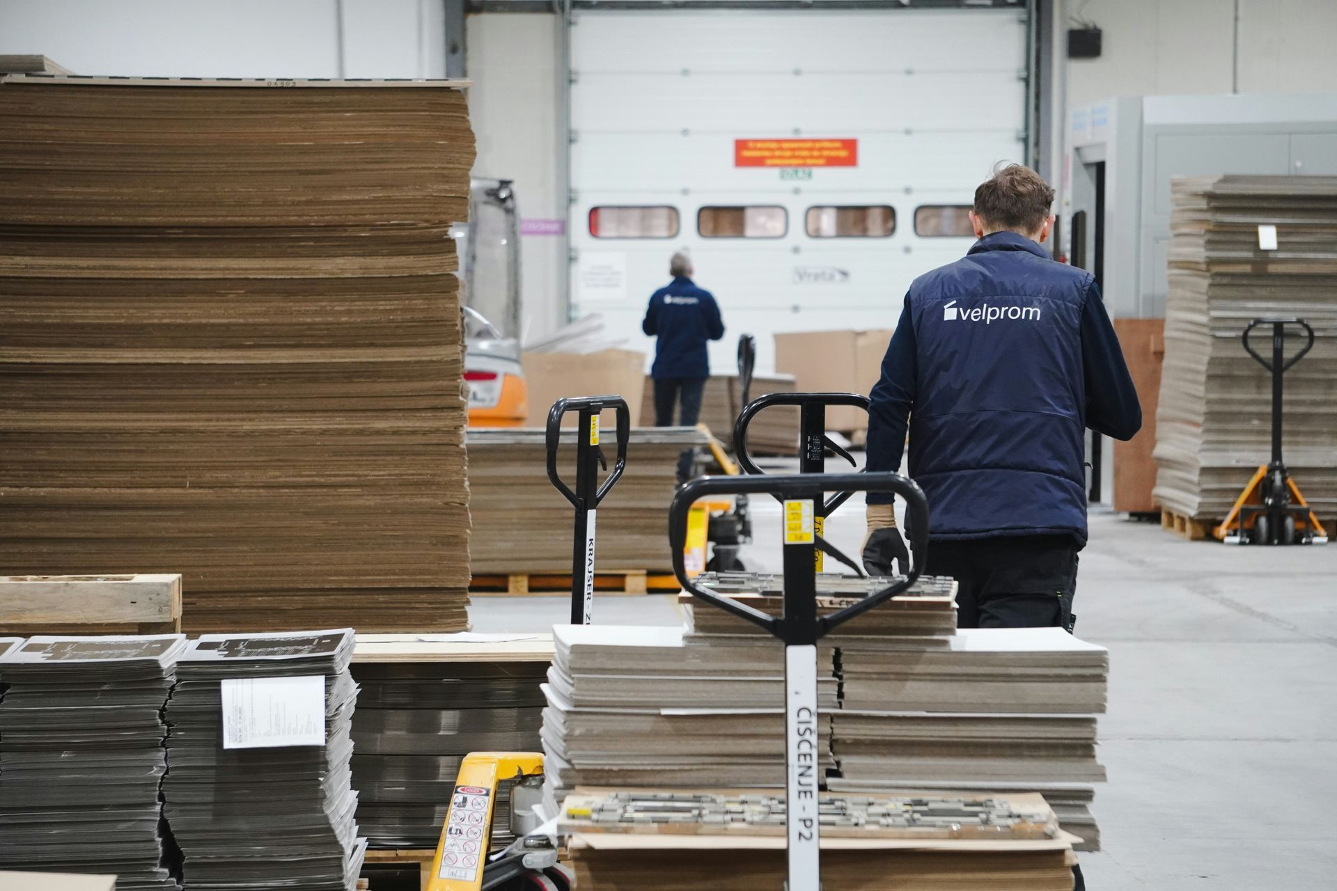 Warehouse workers with pallet jacks near stacked cardboard, loading bay in background.