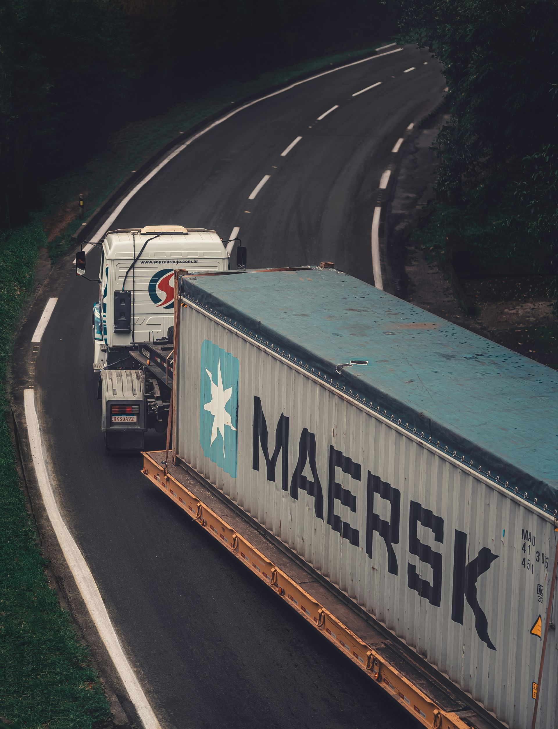 Truck carrying a Maersk shipping container driving on a curved road in a rural setting.