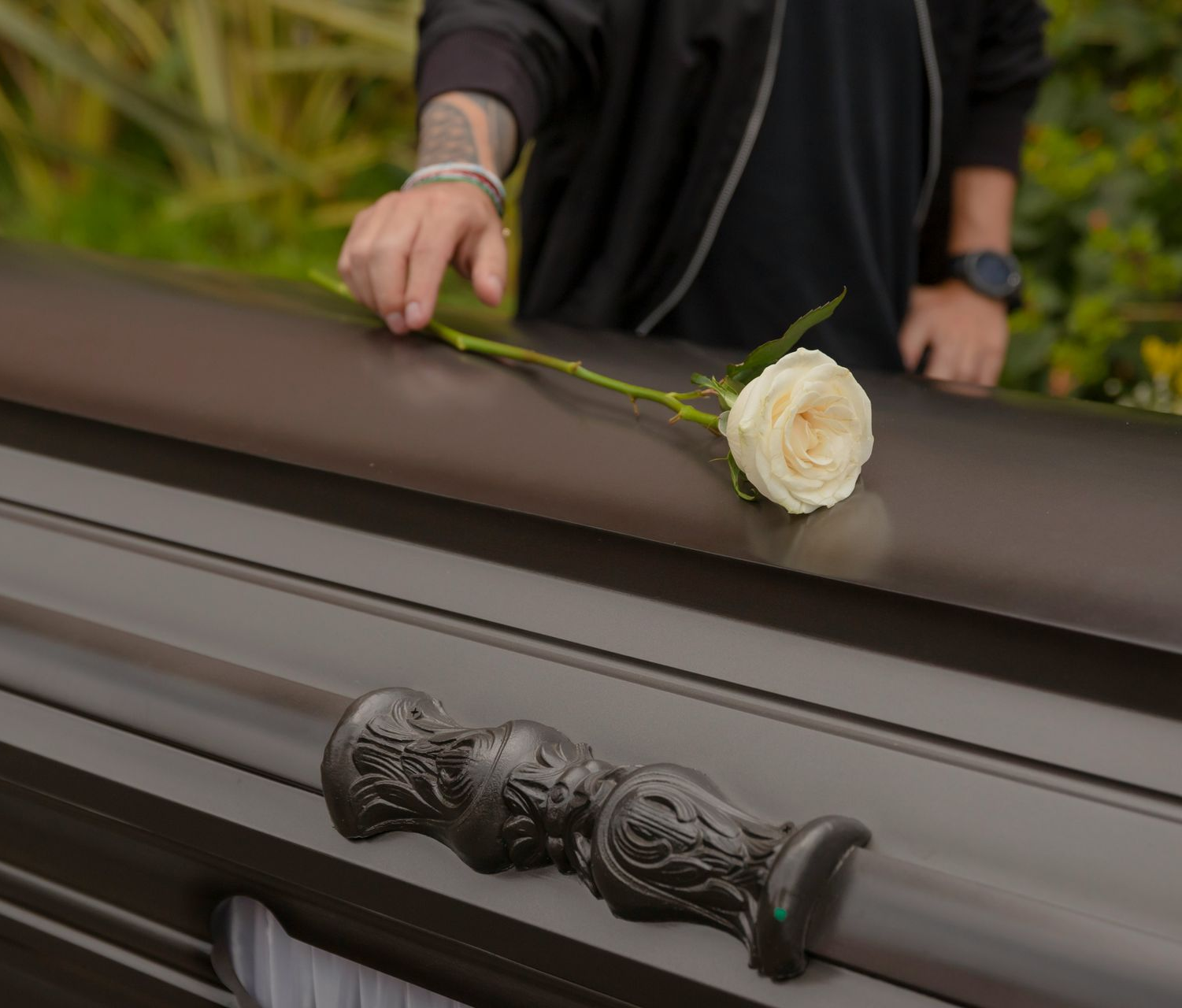 Person's hand with tattoos placing a white rose on a dark coffin, likely at a funeral.