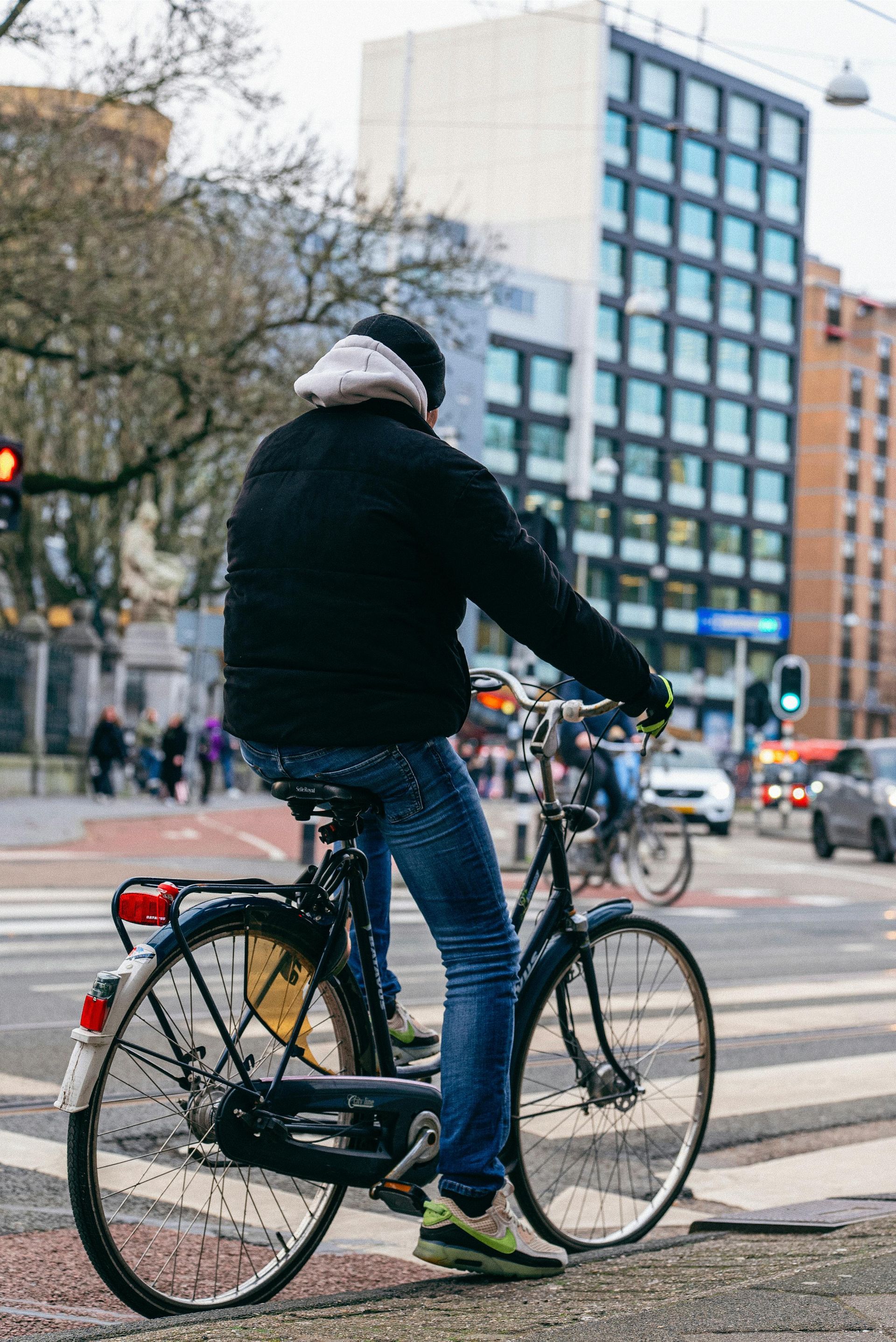 Person on a bicycle at a crosswalk. Black jacket, blue jeans, city street with buildings in background.