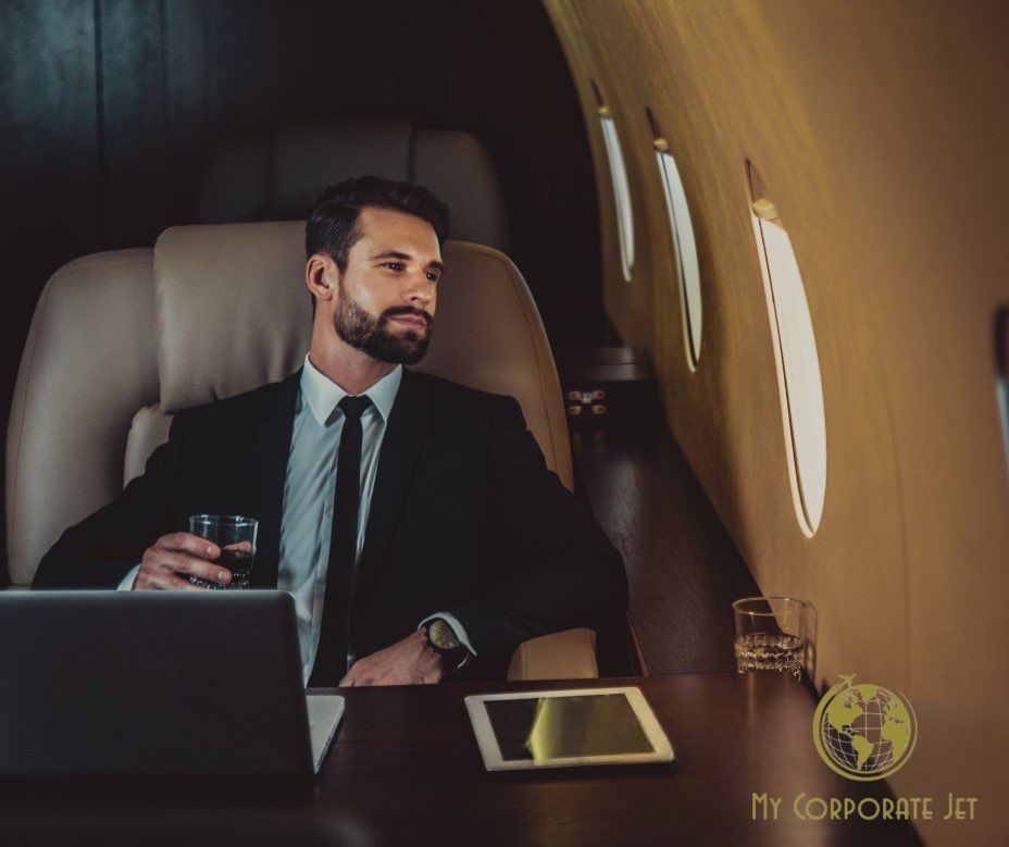 A man in a suit is sitting in an airplane with a laptop and a glass of wine