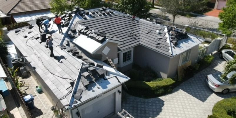 Workers replace roof tiles on a house. The house is a light gray with some old tiles on it.