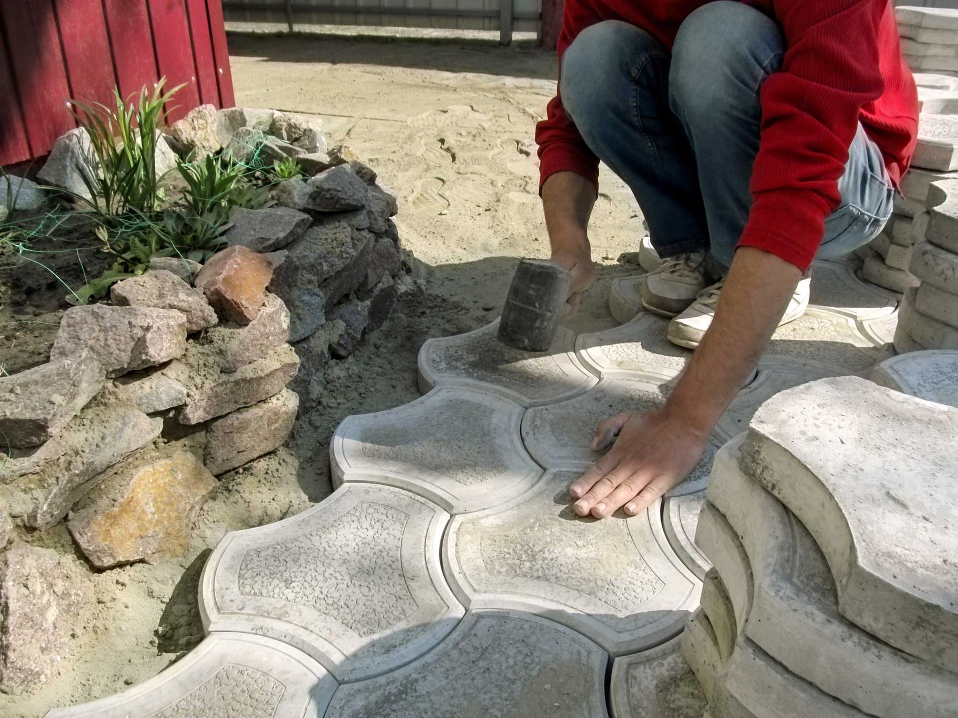A person in a red shirt kneels outdoors, using a mallet to install interlocking grey stone pavers for a garden path.