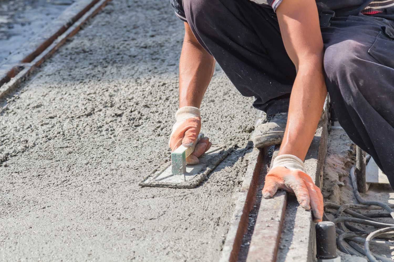 A construction worker uses a hand trowel to smooth wet concrete along the edge of a metal frame.