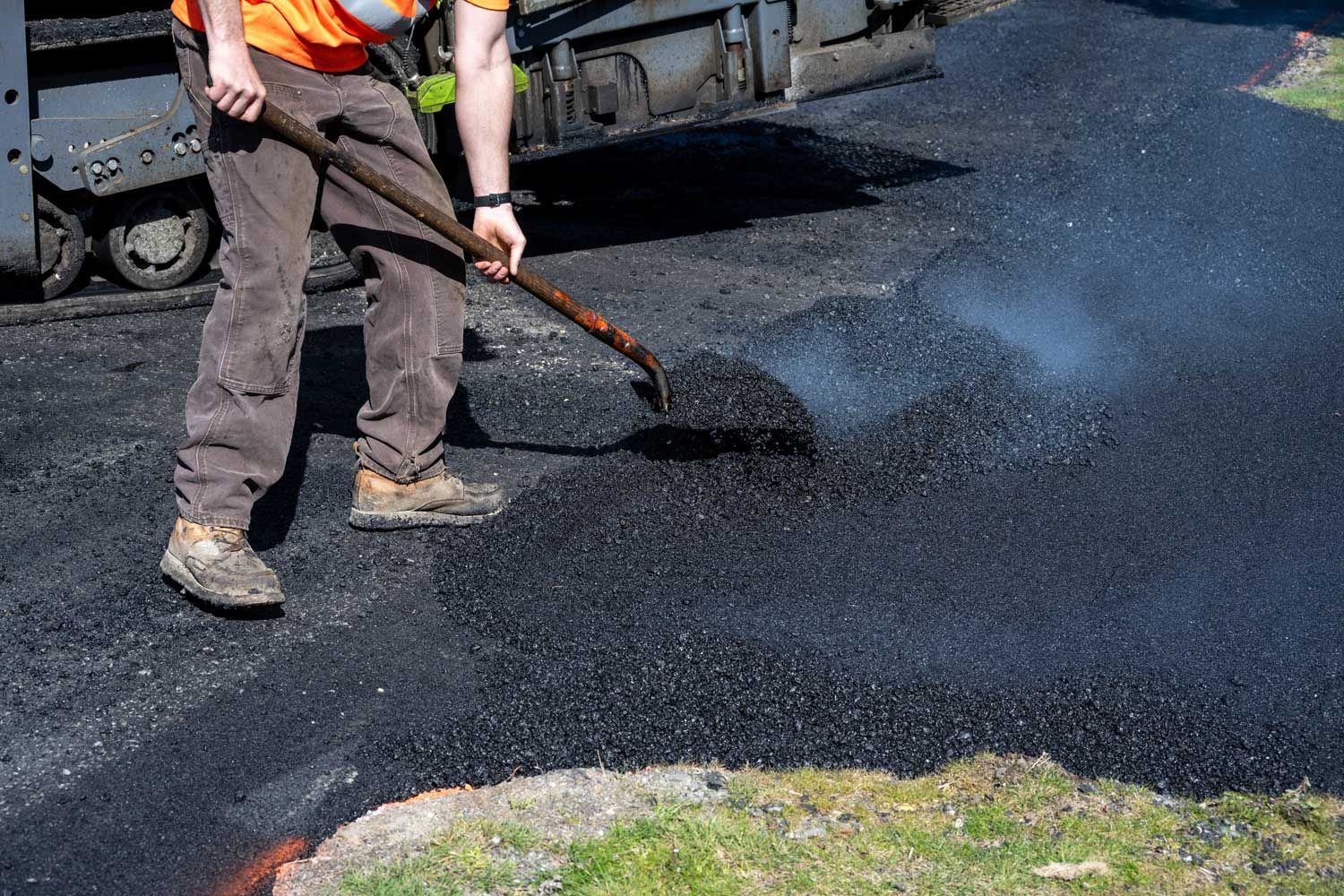 A worker in an orange vest uses a shovel to smooth fresh, steaming black asphalt on a road construction site.