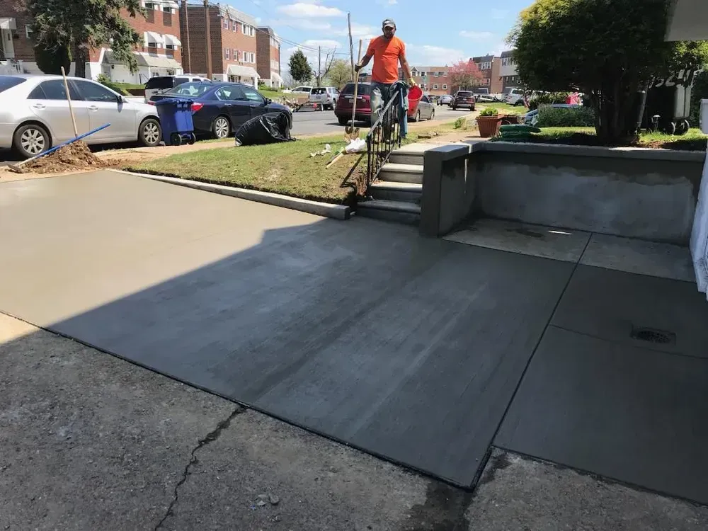 A worker in an orange shirt standing by newly poured concrete steps and a driveway on a sunny day.