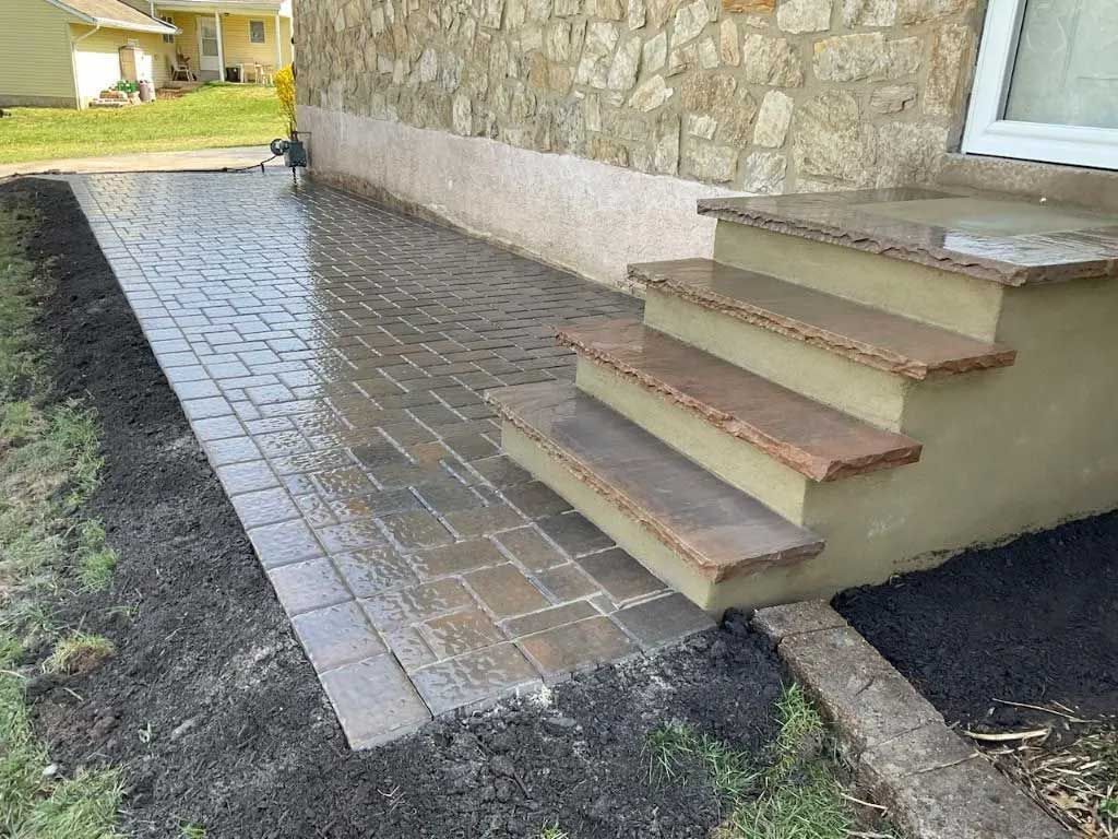 A stone patio leads to three steps with brown stone treads at the base of a textured stone house wall with dark mulch.