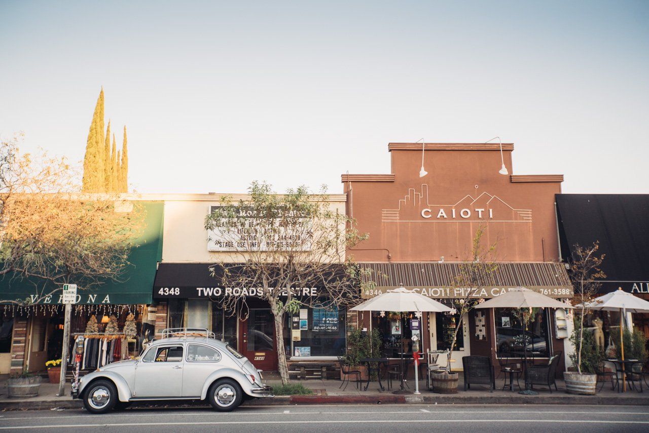 Silver VW Beetle parked in front of shops on a street. Exterior cafe seating, brown buildings, blue sky.