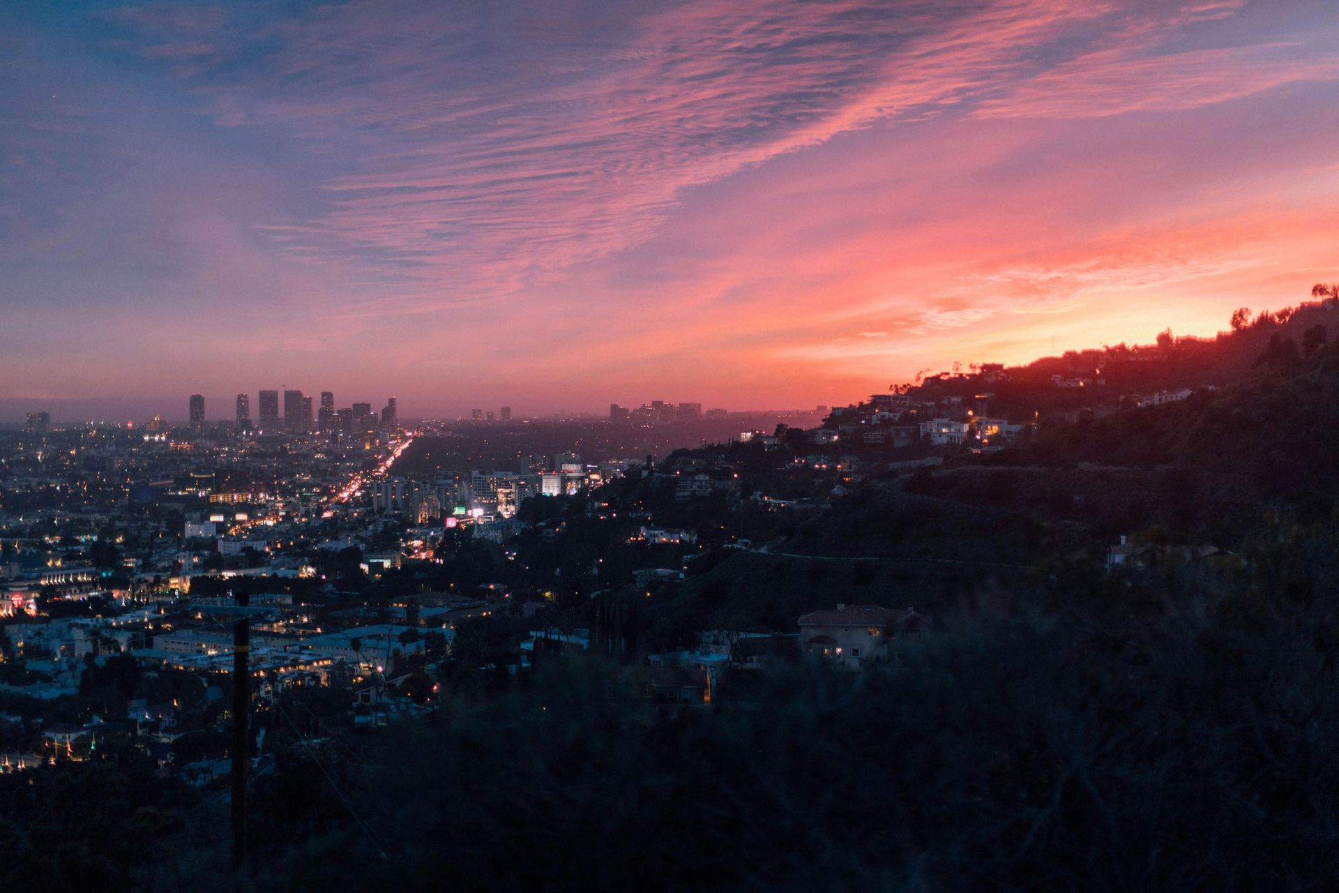 Sunset over Los Angeles, with a bright pink and orange sky. City lights illuminate the buildings and streets below.