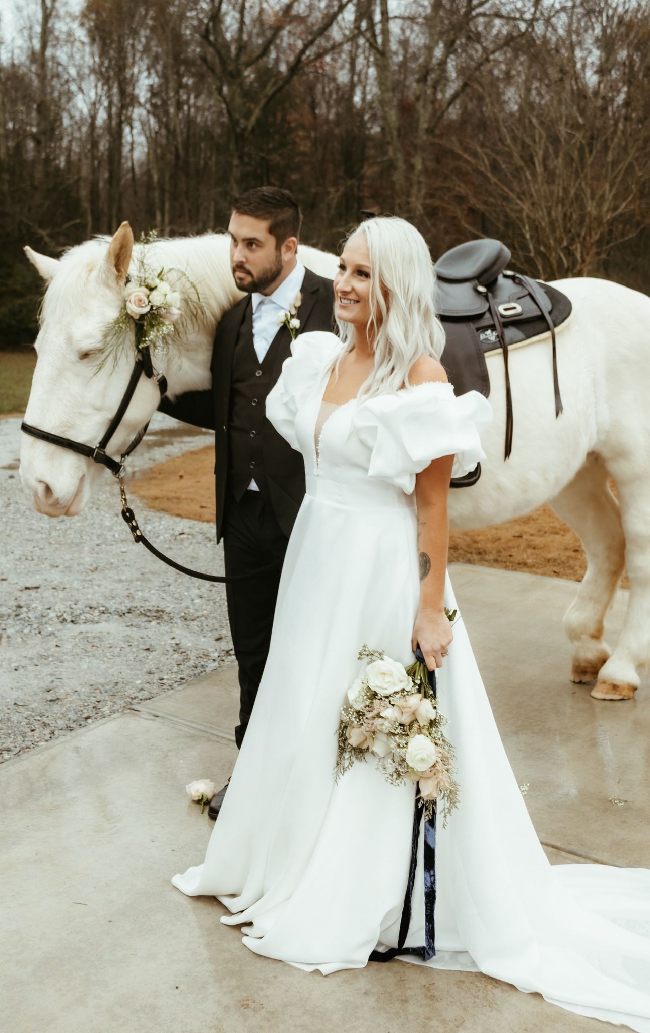 Bride and groom pose with white horse, she in gown holding bouquet, he in suit. Outdoor setting.