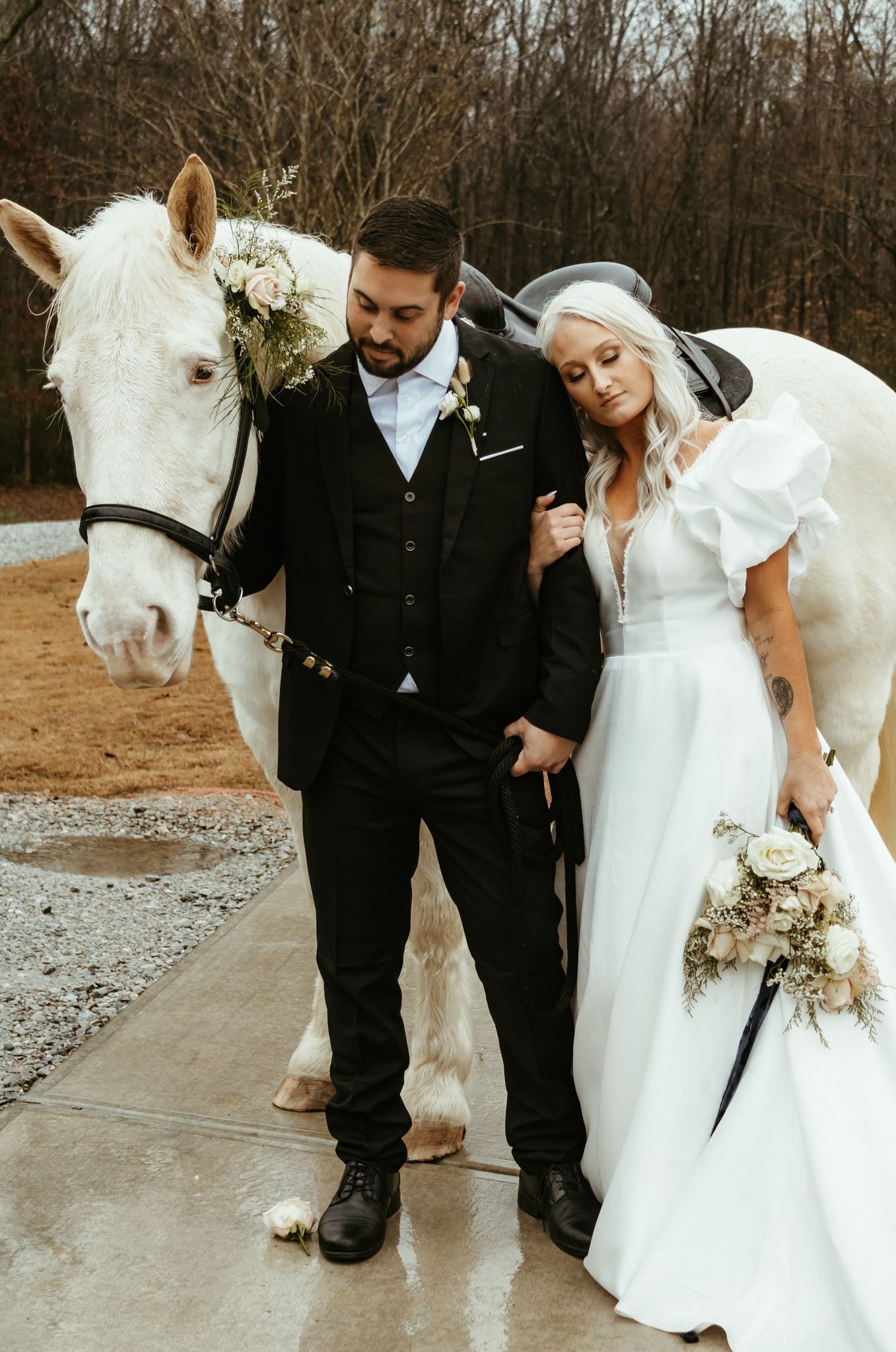 A bride and groom pose with a white horse. The bride wears a white gown, and the groom wears a black suit.