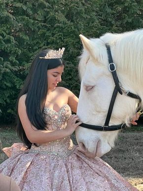 Young woman in a sparkly ball gown and crown petting a white horse; outdoor setting.