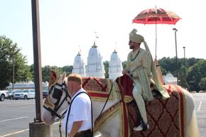 Man in ornate Indian wedding attire rides a white horse, red umbrella overhead, in front of a white temple.