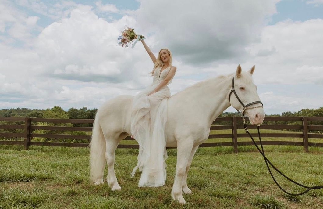 Bride in white dress on a white horse, holding flowers, arm raised, in field with wooden fence.