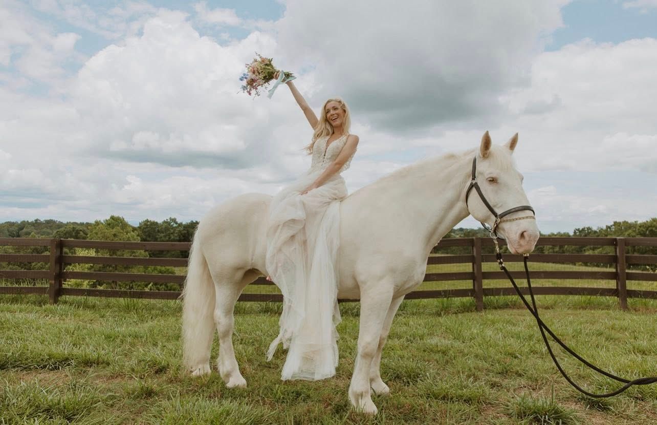 Bride in white dress on a white horse, holding flowers, arm raised, in field with wooden fence.