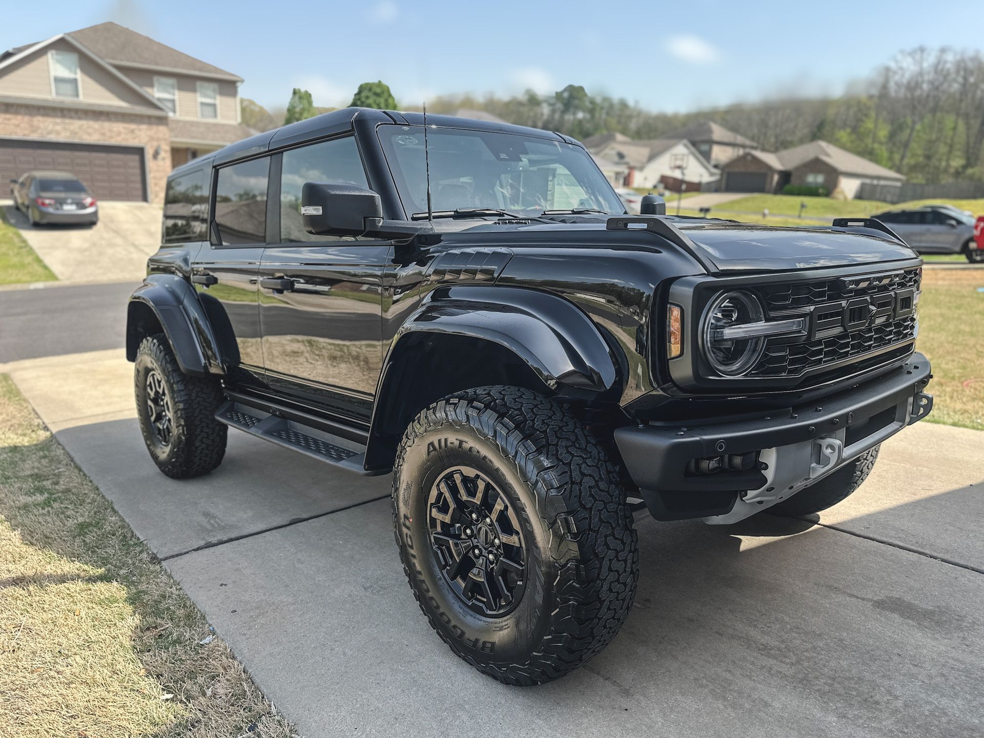 Black Ford Bronco SUV parked on a concrete driveway in front of a house on a sunny day.