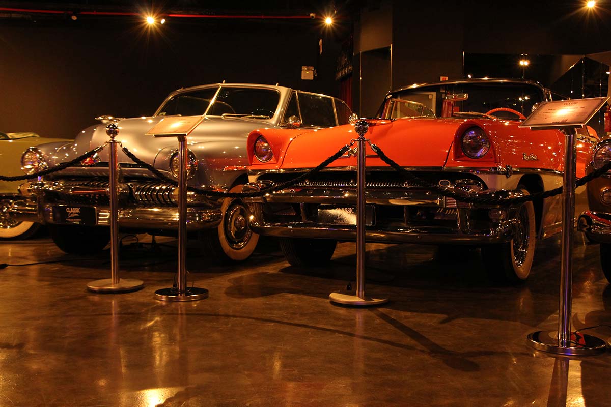 Carros clássicos em exposição, incluindo um Cadillac prata e um Ford laranja, em um ambiente de museu.