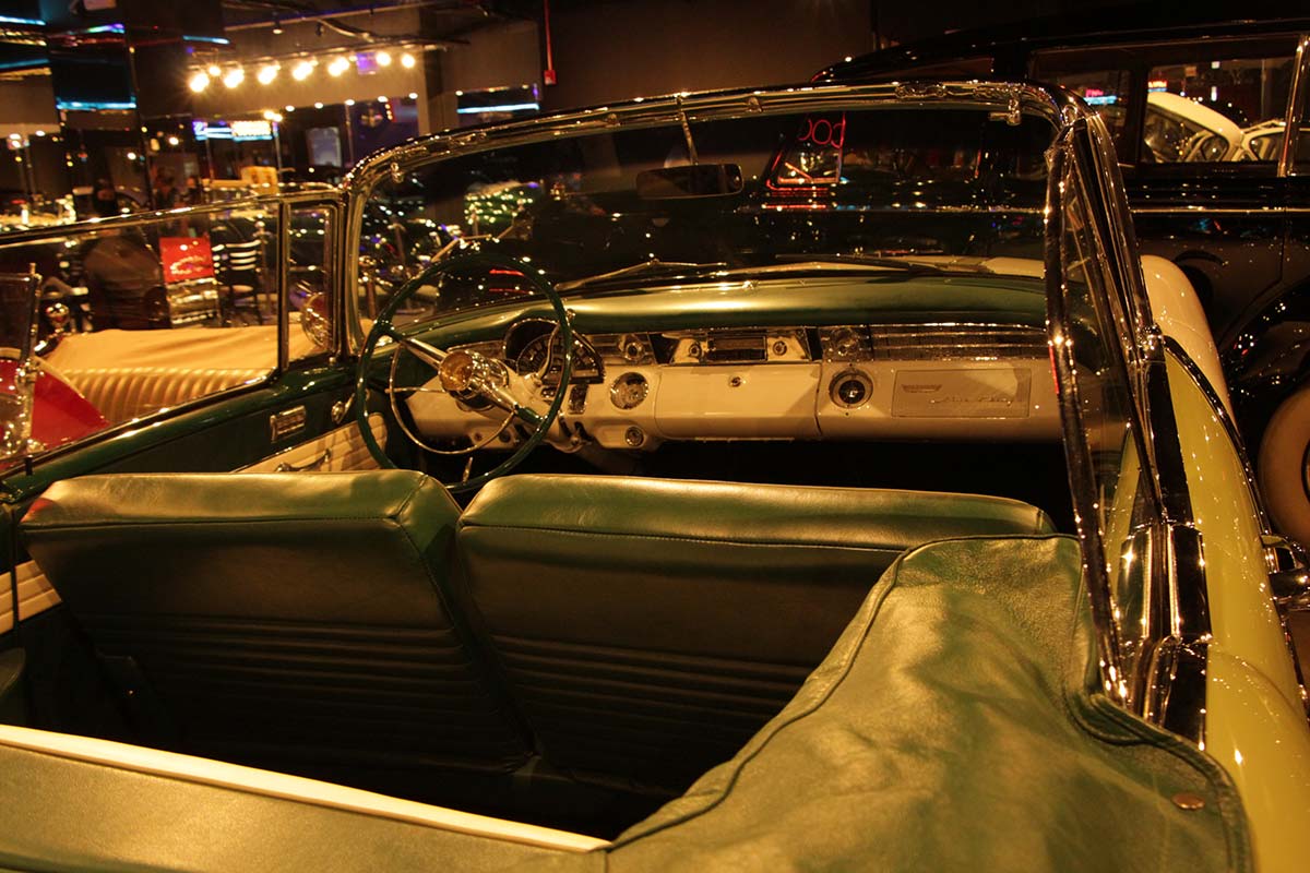 Interior de um carro conversível verde clássico com painel branco e bancos verdes, dentro de um showroom.