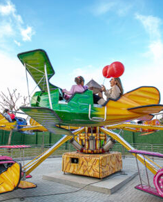 Pessoas andando em um avião verde e amarelo em um parque de diversões, segurando balões vermelhos, sob um céu azul.