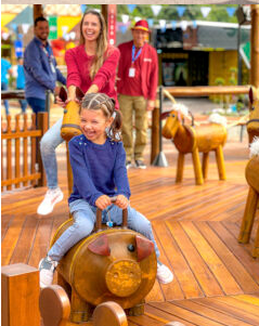 Menina montada em um porco de madeira, sorrindo; mulher no cavalo, homem assistindo em um parque de diversões.