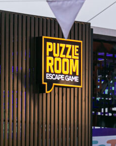 Placa para Puzzle Room Escape Game, preta e amarela contra uma parede vertical de madeira.
