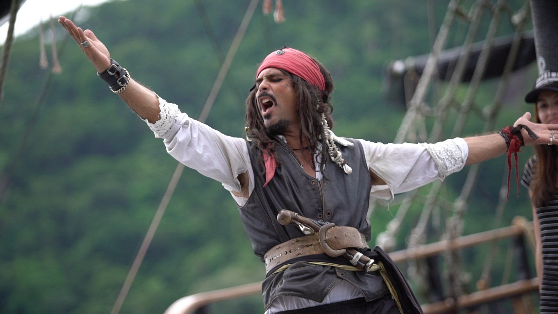 Personagem Jack Sparrow com os braços estendidos, a bordo de um navio, bandana vermelha, boca aberta.