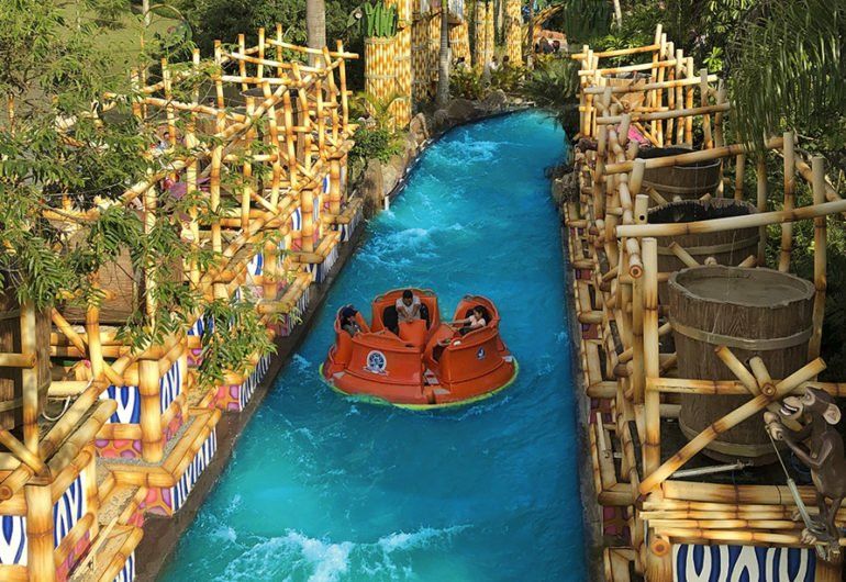 Um passeio aquático em um parque temático. Uma jangada laranja em um rio azul-turquesa, cercada por estruturas decorativas de madeira.