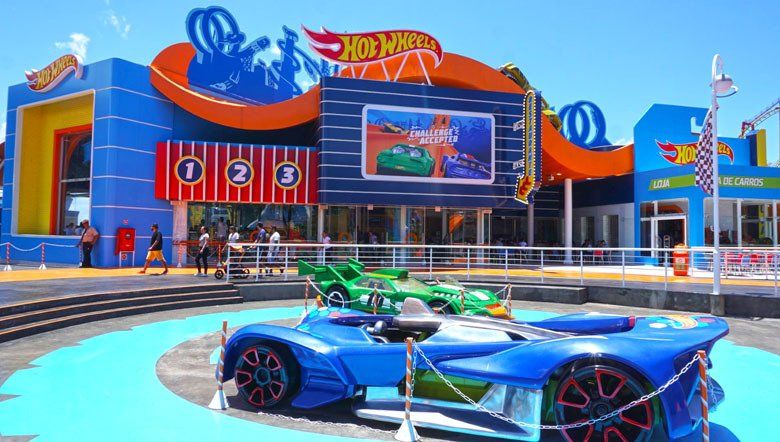Entrada da atração Hot Wheels com carros de corrida coloridos em exposição, prédio azul e laranja.