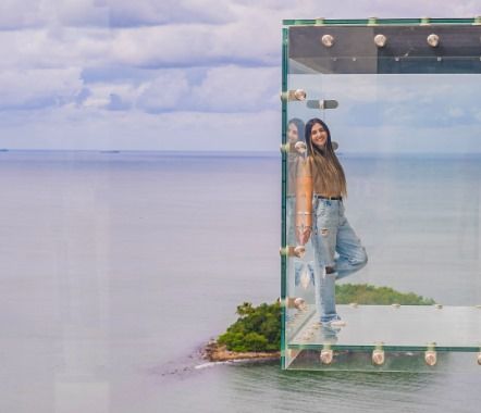 Mulher dentro de uma caixa de vidro com vista para o oceano, sorrindo. Ela veste jeans e uma jaqueta.
