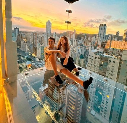 Casal sentado em uma varanda de vidro com vista para a cidade ao pôr do sol.