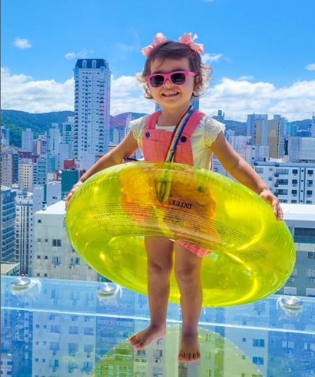 Criança sorrindo com óculos escuros e boia rosa contra o horizonte da cidade.