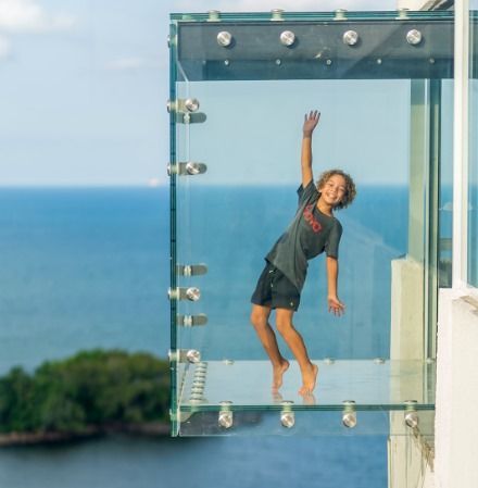 Um menino sorri e acena de uma sacada de vidro com vista para o oceano.