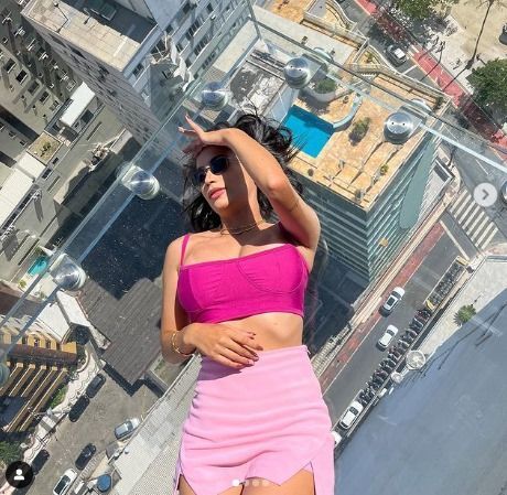 Mulher vestindo top curto e saia rosa, deitada no chão de vidro no alto de uma cidade, bloqueando o sol.