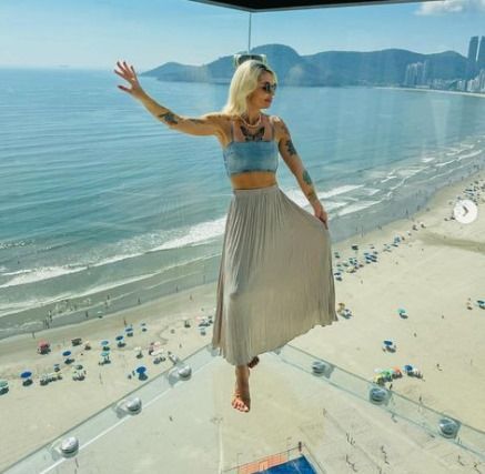 Mulher loira em uma plataforma de vidro sobre uma praia, com os braços erguidos. Oceano e montanhas ao fundo.