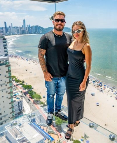 Casal em uma plataforma de vidro com vista para a praia e o horizonte da cidade.