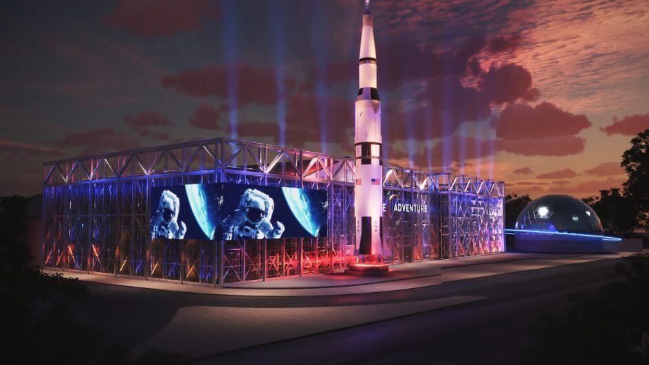 Visão noturna de um edifício com um foguete, iluminado com luzes coloridas, projetando imagens do espaço; uma cúpula de planetário ao lado.