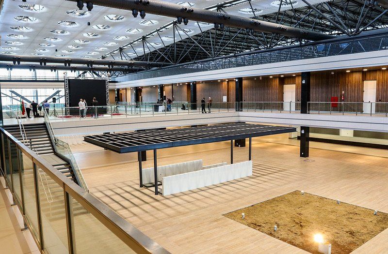 Salão moderno e espaçoso com palco, obra de arte geométrica e passarelas de dois andares, banhado por luz natural.