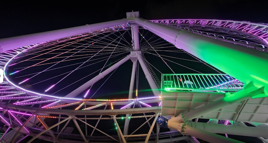 Visão noturna de uma roda-gigante bem iluminada com luzes verdes e roxas contra um céu escuro.
