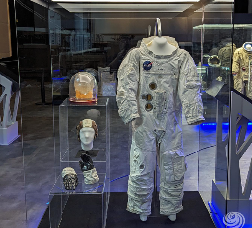 Traje espacial e capacete da Apollo expostos em um museu; traje branco e dourado, manequins, vitrines de vidro.