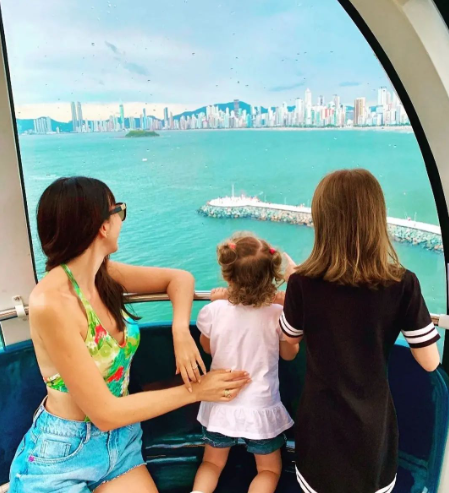 Mulher e duas crianças olhando para a água azul-turquesa e o horizonte da cidade.