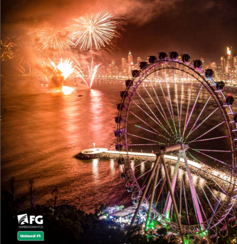 Fogos de artifício explodem sobre uma paisagem urbana e água, com uma roda-gigante iluminada em primeiro plano.