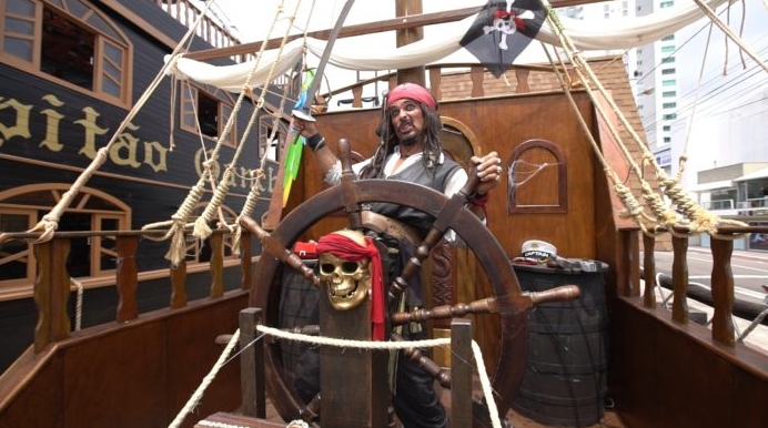 Uma pessoa vestida como Capitão Jack Sparrow em uma réplica de navio pirata, segurando uma espada e sorrindo.