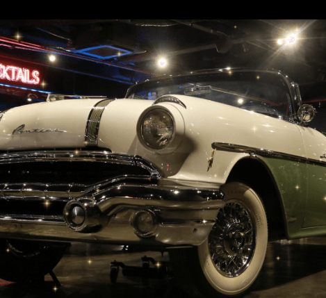 Carro conversível vintage branco e verde com detalhes cromados, iluminado em uma sala com pouca iluminação.