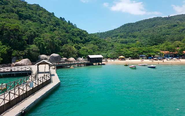 Um píer se estende em águas azul-turquesa em direção a uma praia cercada por montanhas verdejantes sob um céu azul.