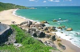 Praia com águas azul-turquesa, faixa de areia, vegetação verde e grandes pedras. Dia ensolarado.